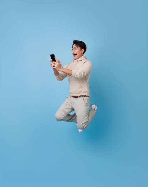 istock Full length handsome Asian man using smartphone he is run and jumping in air isolated on blue background. Young male happy excited and use mobile phone with shopping online concept. 2156899220
