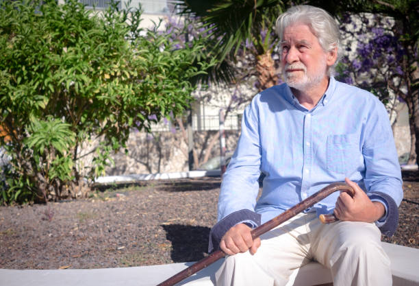 istock Old senior man with hands over the cane sitting in public park with sad expression. Elderly retiree with back pain using walking stick. Tree and flowers on background 2156911439