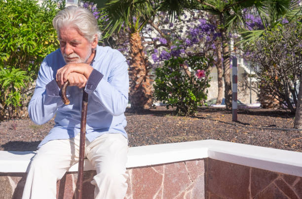 istock Old senior man with hands over the cane sitting in public park with sad expression. Elderly retiree with back pain using walking stick. Tree and flowers on background 2156911514