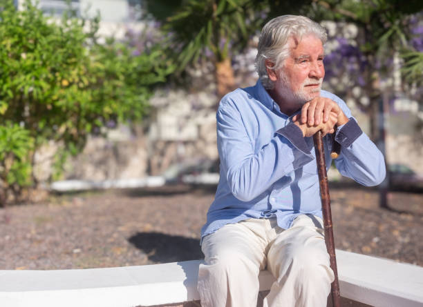 Suffering sad senior bearded man sitting outdoors using a walking pole for legs or back pains. Pensive caucasian male in retirement lifestyle