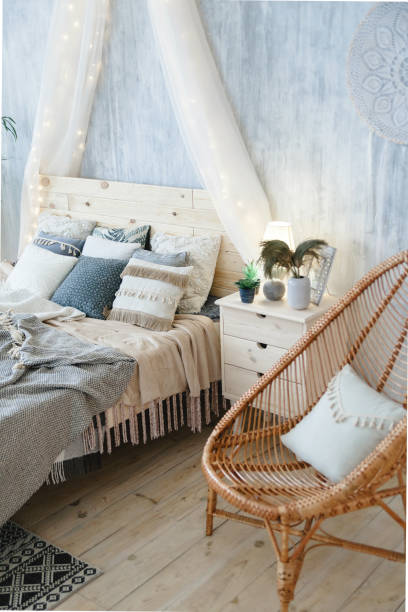 istock Stylish boho bed with wooden headboard, soft blankets, assorted pillows, and light grey walls. Accompanied by wicker chair, macrame wall hanging, bedside table, and decorative vase with dried plants 2156918256