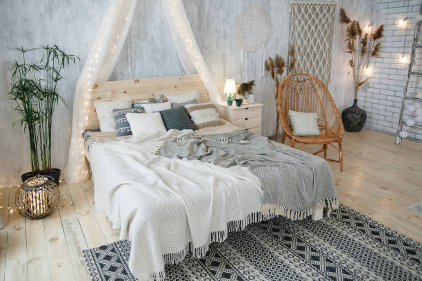 istock Stylish boho bedroom featuring a comfy bed with blankets, wicker chair, green plants, light grey walls, white lights, and bohemian decor elements. Room includes a woven rug and intricate details 2156918817