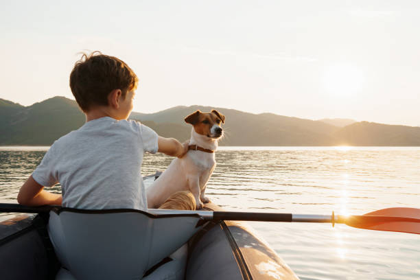 Happy boy with his dog Jack Russell Terrier paddling an inflatable kayak on water mountain lake against backdrop of beautiful orange sunset. Family sports vacation. Lens flare. Pet. Travel concept niño feliz con su perro jack russell terrier remando en un kayak inflable en el lago de la montaña de agua con el telón de fondo de la hermosa puesta de sol naranja. vacaciones deportivas en familia. destello de lente. mascota. concepto de viaje