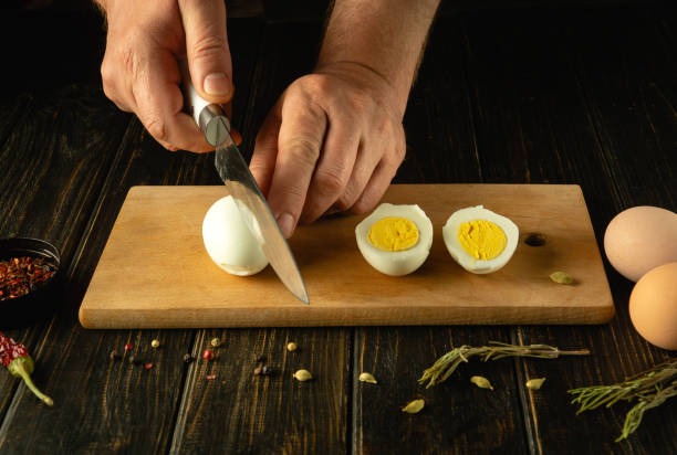istock A man hands cutting a hard-boiled egg in half on a wooden surface next to another hard-boiled egg, already cut, with a yellow yolk in view 2156931643