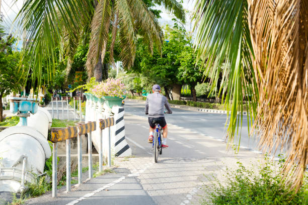 Thai senior man is cyclingg on bicycle lane at Pradit Manutham Road in Bangkok Ramintra. He has passed some trees and is wearing a cycling helmet and s;ports clothing. On street Is no traffic. Rear view of man passing curved part of cycling lane