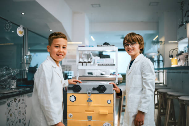 Two young boys wearing lab coats are smiling and standing beside scientific equipment in a modern science laboratory.