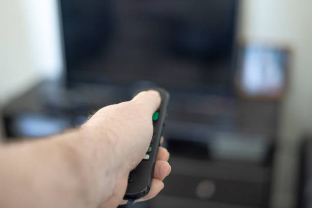 Man pointing television remote control at the TV to turn it on.