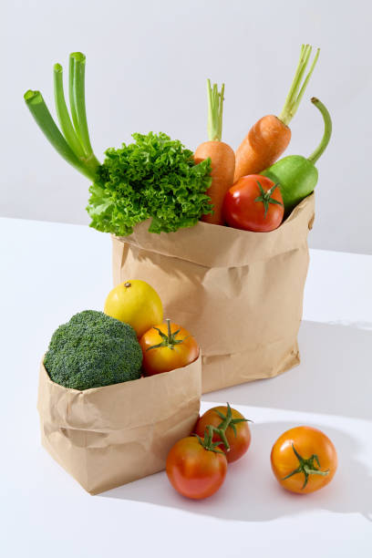 istock Advertising template from high angle shot, two brown paper bag contains fresh vegetable, placed on white table, beside it is some ripe tomatoes. Empty space for designing 2157071550