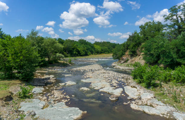 istock Shallow river in a wide stone bed surrounded by a rocky shore and trees. Bright blue sky with clouds 2157073241