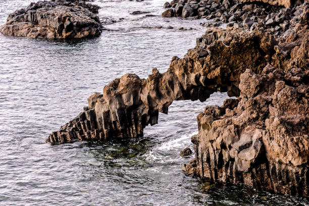 A rocky shoreline with a large archway in the middle. The archway is surrounded by rocks and the water is calm, image