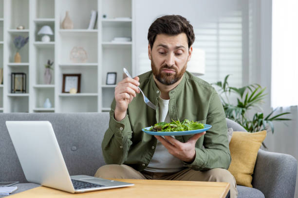 istock Man with a confused expression holding a salad plate while sitting at home with his laptop 2157090111