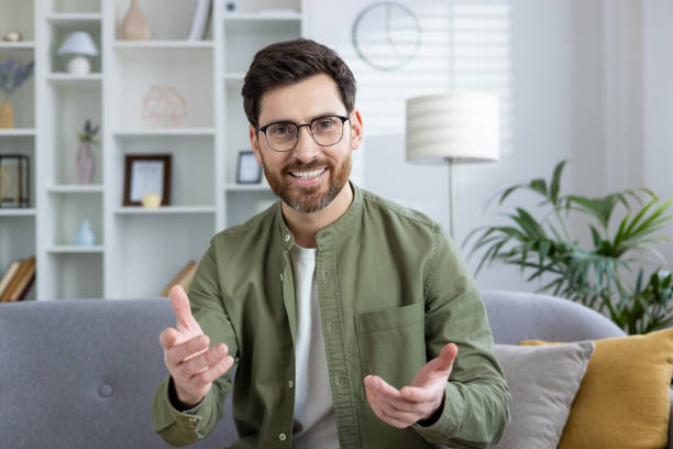 Smiling young man in shorts sitting on sofa at home and talking casually on video call, explaining while gesturing with hands to camera