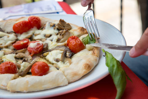 A person is using a fork to slice a pizza. The scene involves food, tableware, ingredients, a recipe, a plate, fast food, kitchen utensils, staple food, produce, and a dish