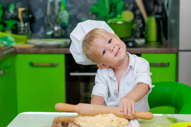 istock happy little boy preparing dough in kitchen at table. there are dough products on table, dressed as chef 2157093332