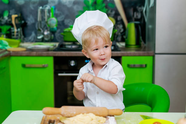 istock happy little boy preparing dough in kitchen at table. there are dough products on table, dressed as chef 2157093334
