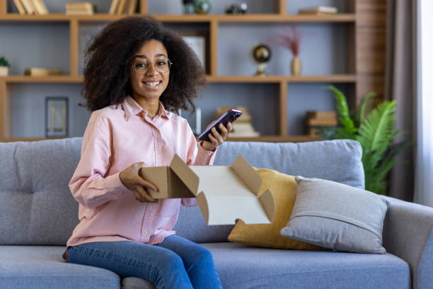 istock Woman opening a package while sitting on a couch and holding a smartphone 2157096917