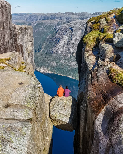 istock Two hikers sit on a rock formation that bridges the gap between two towering cliffs in Norway, enjoying the breathtaking view of the surrounding landscape 2157111033