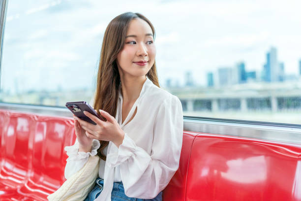 istock Asian woman sitting on a metro skytrain, using her phone and looking away. Background is cityscape 2157113201