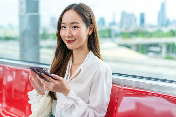 istock Asian woman sitting on a metro skytrain, using her phone and looking away. Background is beautiful cityscape 2157113690