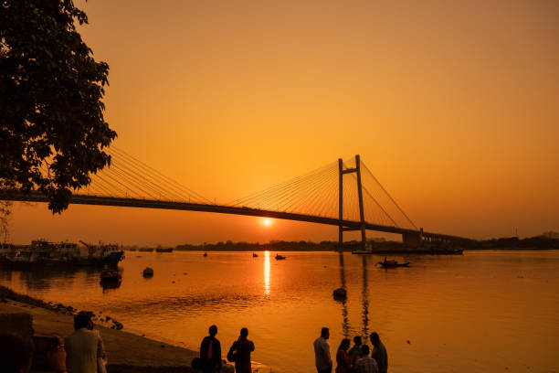 Silhouette people having leisure time at banks of Hooghly river with Vidyasagar Setu in background against sky during sunset