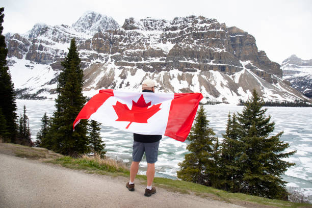 Hiker holds Canadian flag by river and below snowcapped mountains, Bow Lake, Banff National Park