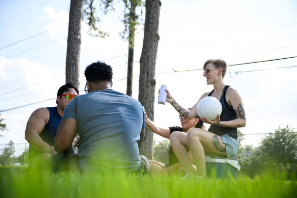 Friends relaxing in park sitting on grass talking together drinking refreshments after volleyball game