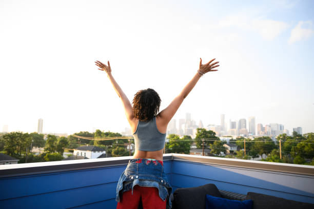 Back view of gen z female on city rooftop extending arms to sky with joy Back view of generation z female on city rooftop extending arms to sky in joy with sunny cityscape in background crop top view stock pictures, royalty-free photos & images