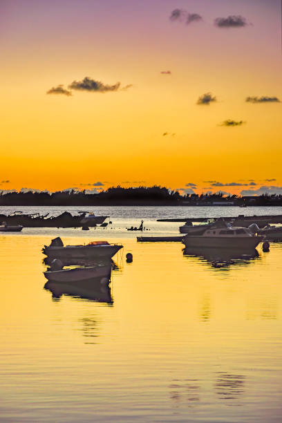 A serene sunset casts lavender and golden hues over a calm body of water where several boats are gently floating. The sky reflects a gradient ranging from a warm yellow to a deepening lavender with sparse wispy clouds, contributing to the tranquil mood of the scene at Spanish Point Bermuda.