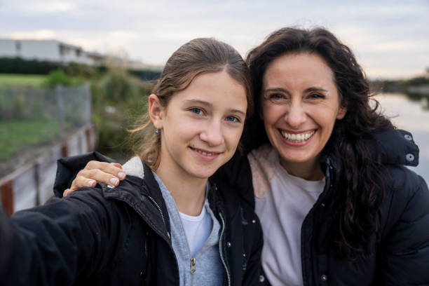 istock Front view of mother and daughter looking at camera and spending time together while sitting on deck by lake 2157154754