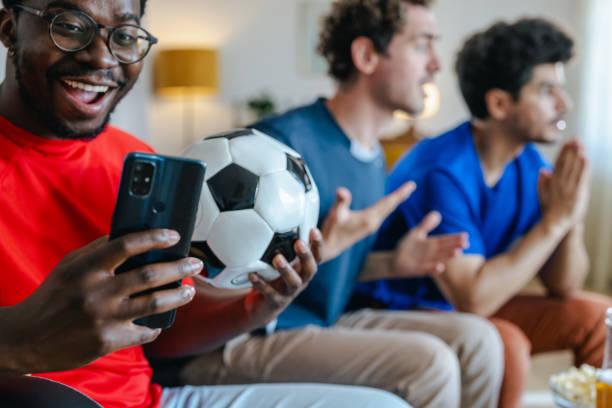 istock Young man using smart phone while watching sports with friends at home 2157157796