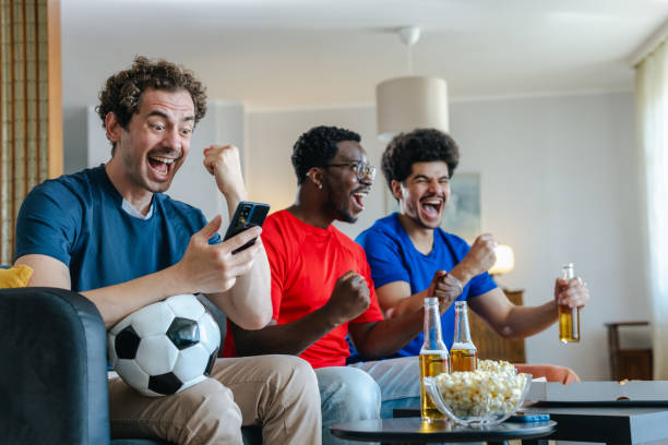 istock Young man using smart phone while watching sports with friends at home 2157157839