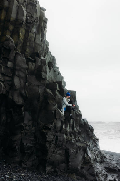 Dramatic view of woman tourist in blue hat sitting on basalt columns and looking at stormy ocean waves in Reynisfjara beach, South Iceland