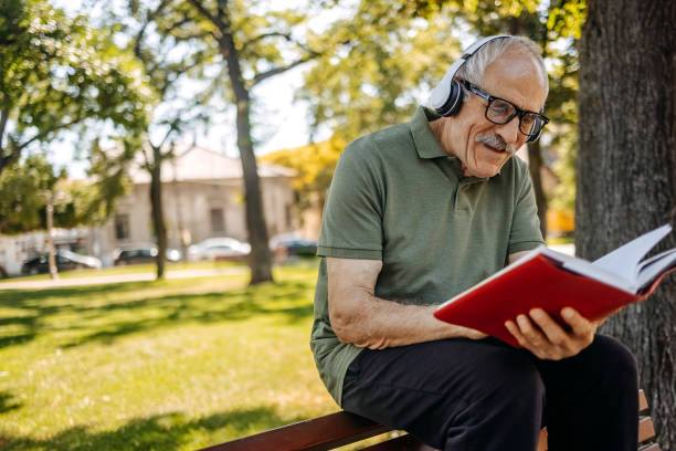 istock Senior man enjoying in hobbies reading a book while sitting in public park on bench 2157176368
