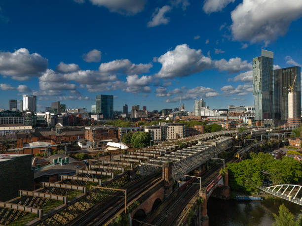 An aerial photograph of an iron railway in Castlefield, Downtown Manchester, Northwest England. The photograph was produced in a warm summer day with intermittent white clouds against a bright blue sky. The Victorian railway leads to Manchester’s modern downtown skyscrapers, which can be seen on the right of the photograph.