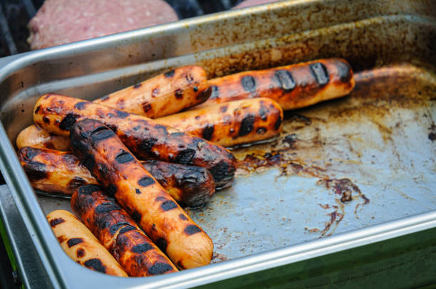 Freshly cooked hot dogs from an outdoor grill wait for their buns in a stainless steel tray at an outdoor barbeque.