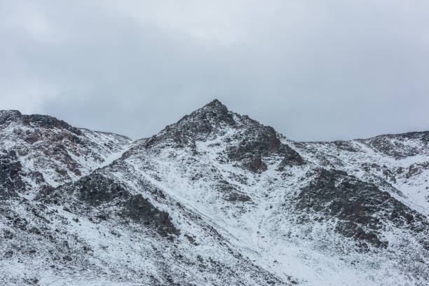 istock Snow-white alpine landscape with large snow-covered mountain range with sharp peaked top. Big snowy rocky ridge with pointy peak in cloudy sky. White snow on rocks. Pointed pinnacle of triangle shape. 2157195024