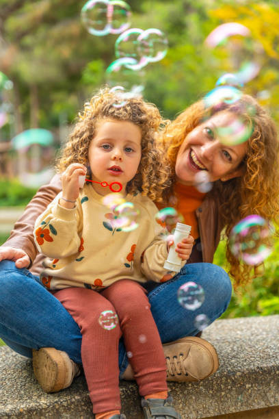 istock Girl and mother sitting on a park blowing bubbles 2157237597