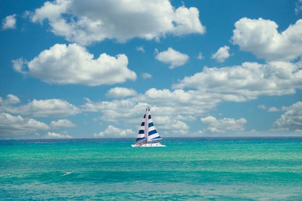 A sailboat with striped sails on a turquoise sea under a blue sky with fluffy clouds.