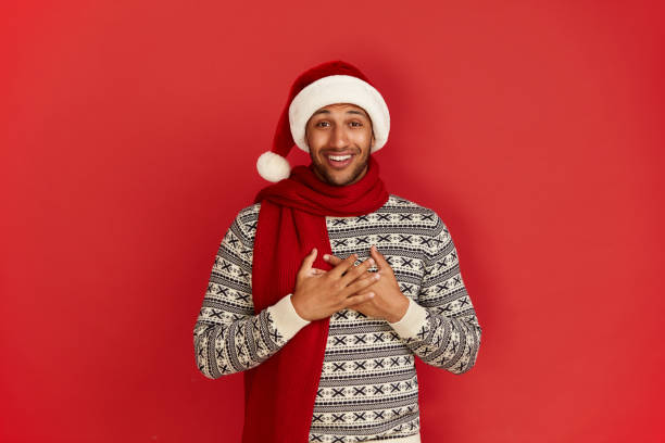 istock Happy Man Smiling Camera. Cheerful Guy in Santa Hat Smiling Sincerely, Holding Hand at Chest, Positive Mood. Indoor Studio Shot, Isolated on Red Background 2157247175