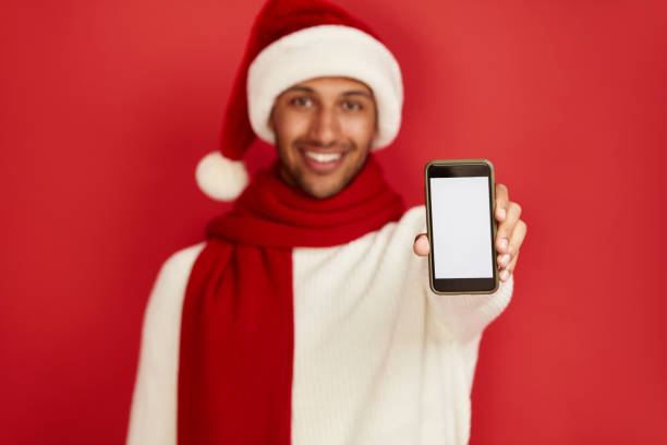 istock Happy Man Using Smartphone. Multiracial Guy in Santa Hat Smiling while Showing Cellphone, Chatting in Social Media, Enjoying Mobile Service. Empty Copy Space for Advertisement, Indoor Studio Shot 2157247200