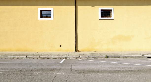 istock Yellow plaster facade with two small barred window and a gutter in the middle. Concrete tiled sidewalk and city street in front. 2157253492