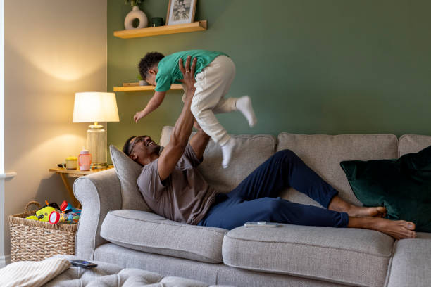 A wide shot of a young boy and his father playing on the sofa in their home in Newcastle-upon-Tyne, North East England. The father is laughing whilst lying down and holding his son high above his head, the young boy's legs blurred from the motion.Videos similar to this scenario available.