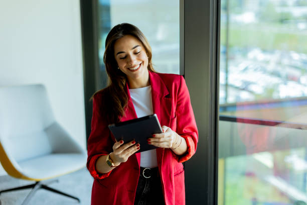 istock Young professional woman in red blazer using tablet by office window on sunny day 2157254227