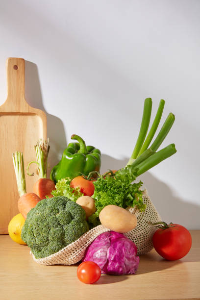 istock Vertical frame photo with white background from frontal shot. In the center, many types of vegetable including tomato, cauliflower, bell pepper, carrot and potato contained in mesh bag on wooden table 2157261484