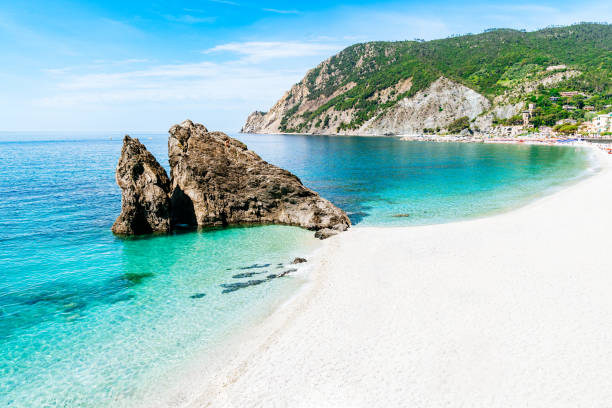 A secluded white sandy beach meets crystal clear shallow waters, leading to deeper turquoise seas. Rocky cliffs rise dramatically on one side, framing the cove, while a Monterosso town nestles against the lush greenery of the hills in the background. Rocky Formation \
