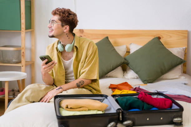 istock Young man holding smartphone and looking away while packing suitcase on bed 2157278845