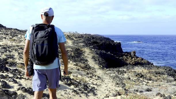 A hiker with a backpack walks along the path on the coast of the volcanic island of Lanzarote in the Canary Islands. Summer tourism activities concept