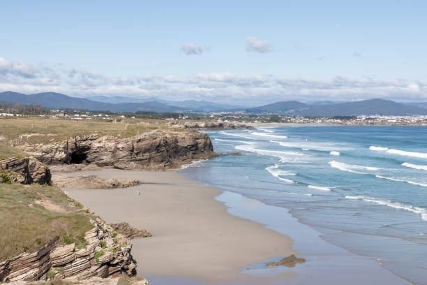 istock Rocky coastline with waves and sandy beach, distant town and mountains under a blue sky. Nature and coastal landscape concept 2157289334