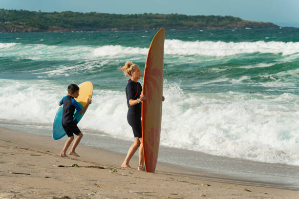 istock Mother and child carrying surfboards on beach 2157307086