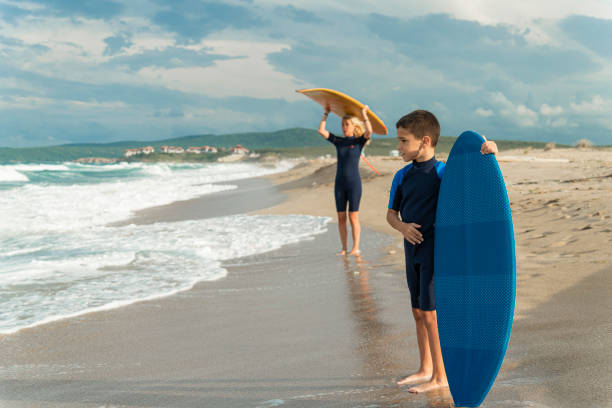 The mother lifts her surfboard demonstrating the technique to her son by the waves, embodying the role of a teacher
